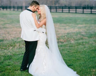 May include: A bride and groom embrace in a field, the bride wearing a white gown with a long veil and the groom wearing a white tuxedo jacket with black pants.