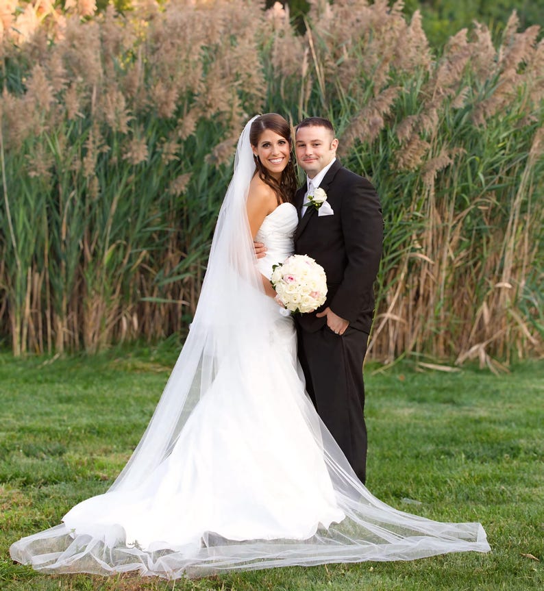May include: A bride and groom pose for a wedding portrait in front of a tall grassy background. The bride is wearing a white wedding dress with a long veil and a bouquet of white flowers. The groom is wearing a black suit and a white shirt with a tie.