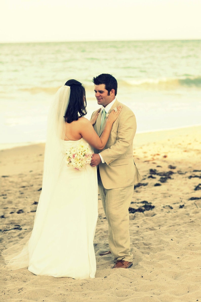 K&ouml;nnte beinhalten: Eine Braut und ein Br&auml;utigam stehen an einem Sandstrand, die Braut tr&auml;gt ein wei&szlig;es Hochzeitskleid und einen Schleier, der Br&auml;utigam einen beigen Anzug. Die Braut h&auml;lt einen Strau&szlig; wei&szlig;er und rosa Blumen.