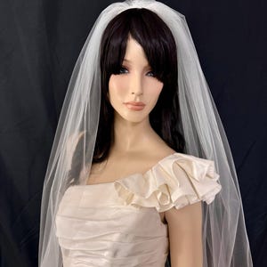 K&ouml;nnte beinhalten: Cremefarbenes Brautkleid mit ger&uuml;schtem &Auml;rmel und passendem Schleier. Das Kleid hat ein Satin-Finish und einen quadratischen Ausschnitt. Der Schleier ist durchsichtig und f&auml;llt &uuml;ber die Schultern. Die Puppe hat dunkles Haar, vor schwarzem Hintergrund.
