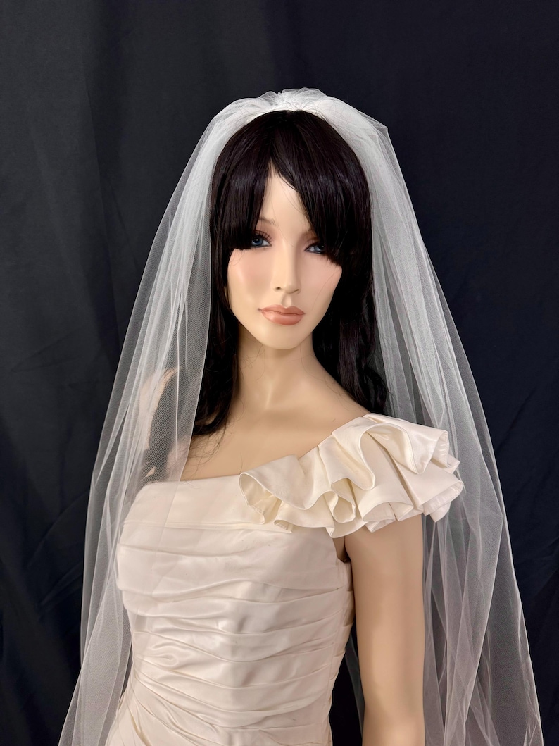 K&ouml;nnte beinhalten: Cremefarbenes Brautkleid mit ger&uuml;schtem &Auml;rmel und passendem Schleier. Das Kleid hat ein Satin-Finish und einen quadratischen Ausschnitt. Der Schleier ist durchsichtig und f&auml;llt &uuml;ber die Schultern. Die Puppe hat dunkles Haar, vor schwarzem Hintergrund.