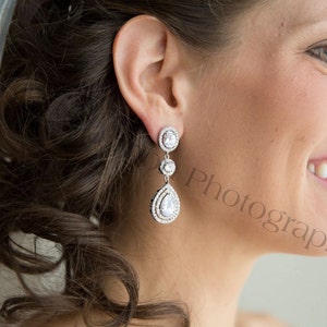 May include: Elegant, three-tiered drop earrings with a sparkling, clear gemstone design. The earrings feature a round top, a smaller round center, and a larger pear-shaped bottom. The earrings are set in a silver-colored metal and are worn by a person with dark hair.