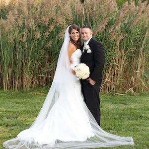 May include: A bride and groom pose for a wedding portrait in front of a tall grassy field. The bride is wearing a white wedding dress with a long veil and a bouquet of white and pink flowers. The groom is wearing a black suit and a white shirt with a tie.