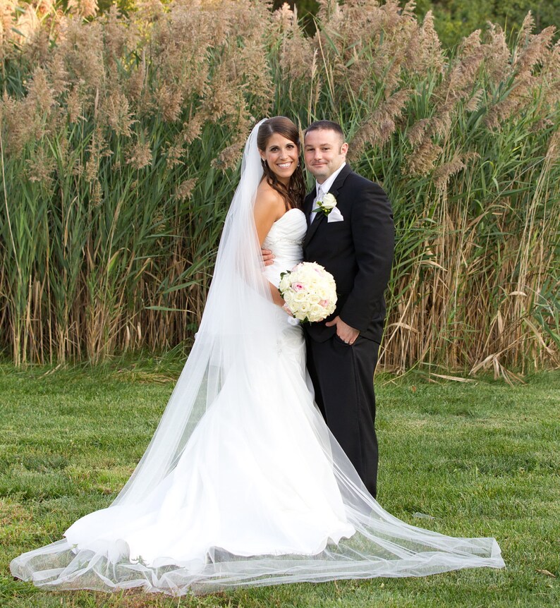 May include: A bride and groom pose for a wedding portrait in front of a tall grassy field. The bride is wearing a white wedding dress with a long veil and a bouquet of white and pink flowers. The groom is wearing a black suit and a white shirt with a tie.