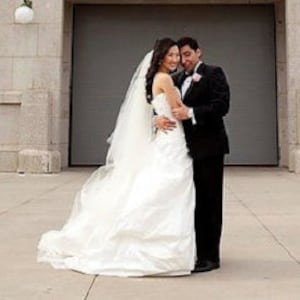 May include: A bride and groom pose for a wedding photo in front of a large gray door. The bride is wearing a white strapless wedding dress with a long veil. The groom is wearing a black tuxedo with a white bow tie.