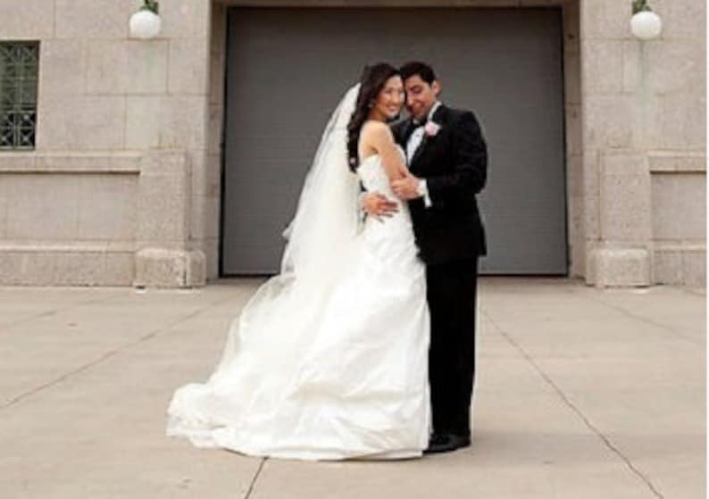 May include: A bride and groom pose for a wedding photo in front of a large gray door. The bride is wearing a white strapless wedding dress with a long veil. The groom is wearing a black tuxedo with a white bow tie.
