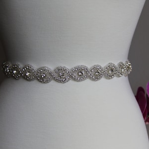 May include: A silver bridal sash with a delicate design of clear rhinestones. The sash is shown on a white dress form.