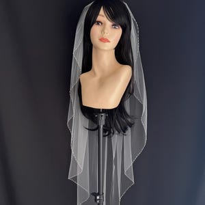 May include: A white bridal veil with a beaded edge. The veil is draped over a mannequin head with long black hair.