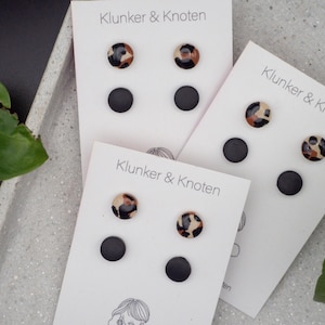 Könnte beinhalten: Mehrere Paare Ohrstecker auf weißen Karten. Jede Karte hat zwei Ohrringe: einen mit einem schwarz-braunen Fleckenmuster und einen einfarbigen schwarzen Ohrring. Die Karten sind mit "Klunker & Knoten" beschriftet.