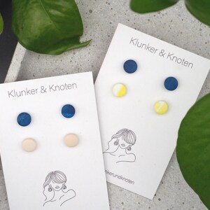 Könnte beinhalten: Zwei Sets Ohrstecker auf weißen Karten mit dem Text "Klunker & Knoten". Ein Set hat marineblaue und hellrosa Ohrstecker, das andere marineblaue und gelbe Ohrstecker. Auf den Karten befindet sich eine Strichzeichnung einer Frau.