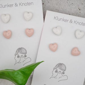 Könnte beinhalten: Zwei weiße Präsentationskarten mit der Aufschrift Klunker & Knoten, die jeweils zwei Paar herzförmige Ohrstecker enthalten. Ein Paar ist weiß und das andere zartrosa. Jede Karte zeigt eine schwarze Strichzeichnung einer Person.