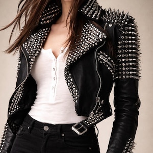 Puede incluir: Chaqueta de cuero negra adornada con tachuelas y púas metálicas plateadas. La chaqueta tiene cierre de cremallera y cinturón. La modelo lleva una camiseta blanca y vaqueros negros.