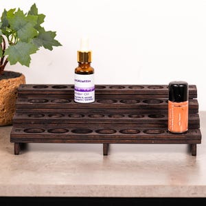 May include: A dark brown wooden tiered display stand with multiple circular cutouts for small bottles. One amber glass bottle with a gold dropper cap and one orange bottle with a black cap are placed on the stand's surface.