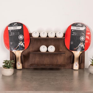 May include: A wooden wall-mounted rack with two red and black ping pong paddles and six white ping pong balls. The paddles have the text "PRO 1000" on them.