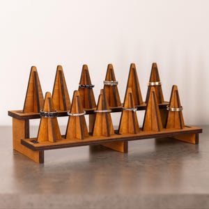 May include: A two-tiered wooden ring display stand featuring twelve triangular cones arranged in two rows. The stand is constructed from light brown wood with dark brown edges, designed to hold rings on each individual cone-shaped post.