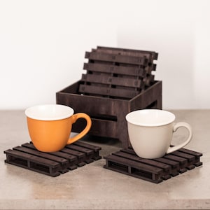 May include: A set of dark brown wooden coasters designed to look like miniature shipping pallets. Two coasters are placed flat, each holding a ceramic mug, one orange and one off-white. A matching wooden crate holds additional stacked coasters.
