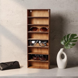 Op de afbeelding: Een houten zonnebrillen organizer met meerdere planken, met drie zonnebrillen. Een zwarte zonnebril etui staat links. Een witte vaas met een groen blad staat rechts. De organizer is gemaakt van lichtbruin hout.