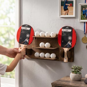 Könnte beinhalten: Ein wandmontiertes Regal aus dunkelbraunem Holz mit zwei Pro 1000 Tischtennisschlägern mit roten und schwarzen Belägen sowie neun weißen Tischtennisbällen auf drei horizontalen Ablagen mit seitlichen Halterungen für die Schlägergriffe.
