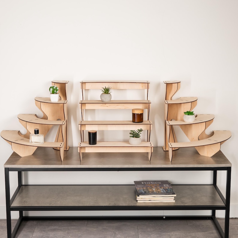 May include: A set of three light-coloured wooden display stands with a smooth finish. The central unit features four tiered shelves, while the two side units have a curved, stepped design. Small potted plants and cylindrical containers sit on the shelves.