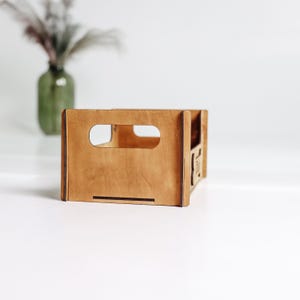 May include: A rectangular wooden crate with a light brown finish, featuring a cutout handle on the front panel. The structure is assembled with visible joints and has an open top, providing a clear view of the interior space and the smooth surface.
