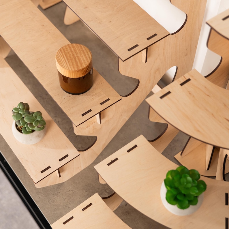 May include: A tiered light-coloured wooden display stand with multiple curved shelves featuring dark rectangular slot joints. The stand holds a small brown jar with a wooden lid and two small green succulent plants in white rounded pots.