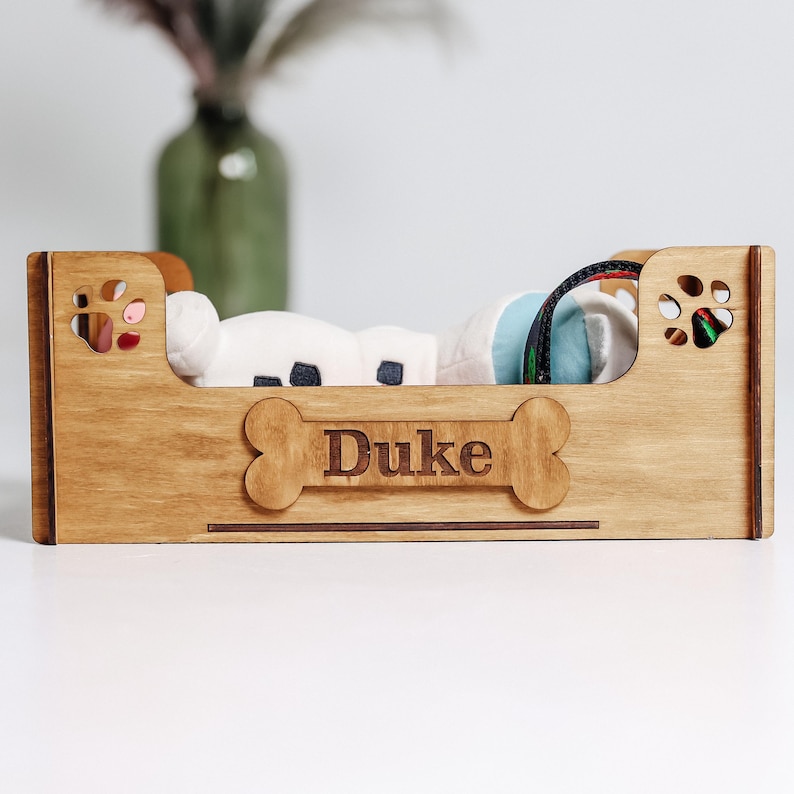 May include: A light brown wooden pet bed featuring a bone-shaped cutout on the front panel with the name Duke engraved in the center. The side panels include paw print cutouts, and the structure holds soft white and blue fabric items inside.