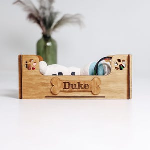 May include: A wooden dog bed with the name "Duke" engraved on the front. The bed is filled with a white and black plush toy, a blue and white toy, and a black and green toy. The sides of the bed have paw-shaped cutouts.