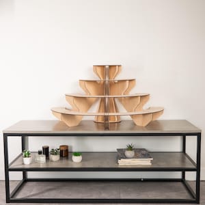 May include: A tiered light-coloured wooden display stand with four levels, featuring a curved, semi-circular design. The stand is constructed from interlocking panels and is positioned on a grey surface with a black metal frame base.