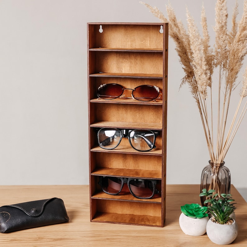 May include: A vertical wooden display rack with eight open shelves, featuring three pairs of sunglasses resting on the middle shelves. The rack has a natural light brown wood grain finish and two circular mounting holes at the top edge.