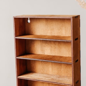May include: A rectangular wooden wall shelf featuring four open storage levels with a natural brown wood grain finish. The structure is constructed with visible interlocking joints on the side panels and includes a circular keyhole cutout.