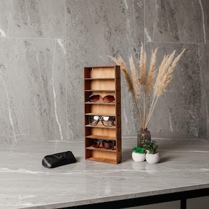 May include: A vertical wooden display rack with seven shelves, holding three pairs of sunglasses on the middle shelves. A black glasses case sits to the left, and a glass vase with dried pampas grass and two small potted succulents sit to the right.