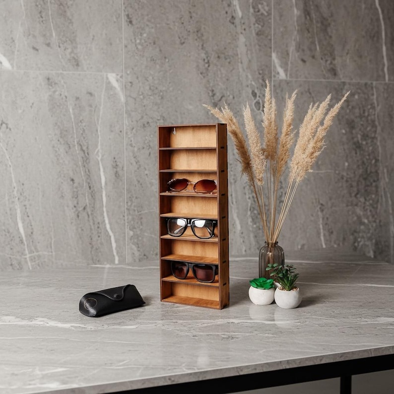 May include: A vertical wooden display rack with seven shelves, holding three pairs of sunglasses on the middle shelves. A black glasses case sits to the left, and a glass vase with dried pampas grass and two small potted succulents sit to the right.