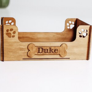 May include: A light brown wooden pet bed frame with a rectangular base and four sides. The front panel features a bone-shaped cutout with the name Duke engraved in the center. Each corner panel includes a laser-cut paw print design.