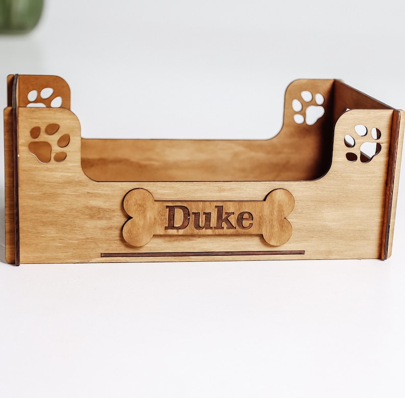 May include: A light brown wooden pet bed frame with a rectangular base and four sides. The front panel features a bone-shaped cutout with the name Duke engraved in the center. Each corner panel includes a laser-cut paw print design.
