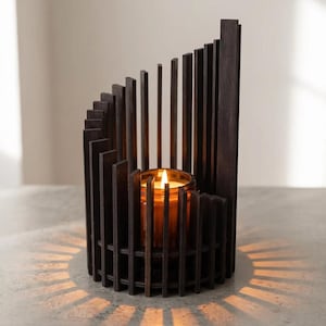 May include: A dark brown wooden candle holder featuring a series of vertical slats arranged in a spiral, ascending height pattern. A glass jar containing a lit candle with a warm orange flame sits in the centre, casting radial light patterns.