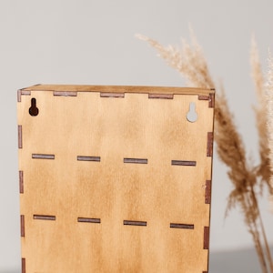 May include: A rectangular light brown wooden wall-mounted display case featuring visible finger joint construction along the edges. The back panel includes two pre-cut keyhole slots at the top for mounting purposes and horizontal slots.