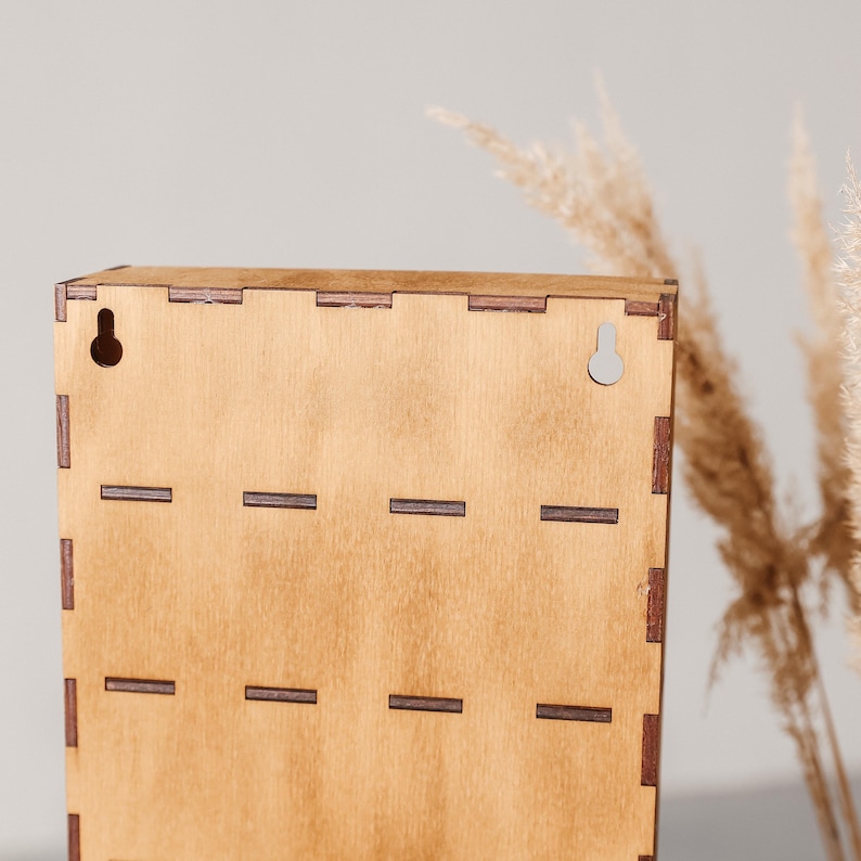 May include: A rectangular light brown wooden wall-mounted display case featuring visible finger joint construction along the edges. The back panel includes two pre-cut keyhole slots at the top for mounting purposes and horizontal slots.