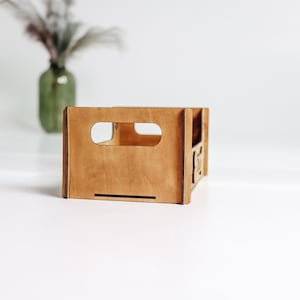 May include: A rectangular wooden crate with a light brown finish, featuring a cutout handle on the front panel. The structure is assembled with visible joints at the corners, and the side panel displays a small, partially visible laser-cut design.
