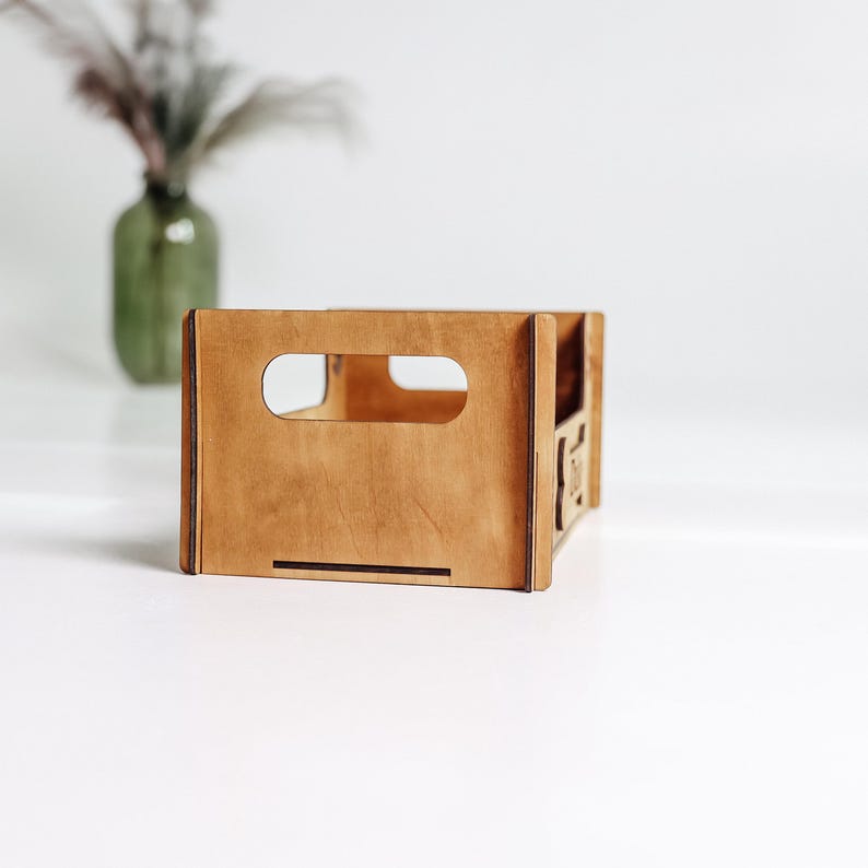 May include: A rectangular wooden crate with a light brown finish, featuring a cutout handle on the front panel. The structure is assembled with visible joints at the corners, and the side panel displays a small, partially visible laser-cut design.