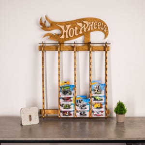 May include: Wooden Hot Wheels display stand with a sign that says "Hot Wheels". The stand has four rows of slots to hold Hot Wheels cars in their packaging. There are several Hot Wheels cars in their packaging on the stand.