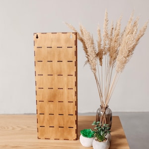 May include: A rectangular light brown wooden wall display board featuring a grid of horizontal slots for organising items. The board has two keyhole mounting holes at the top and a notched edge design along the sides, standing upright on a surface.