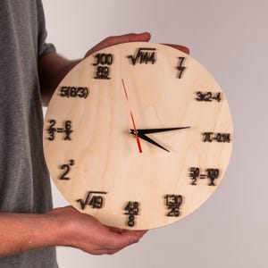 Puede incluir: Un reloj redondo de madera con números y ecuaciones negras en lugar de números tradicionales. Las manecillas del reloj son negras, con un segundero rojo. El reloj se sostiene frente a un fondo neutro.