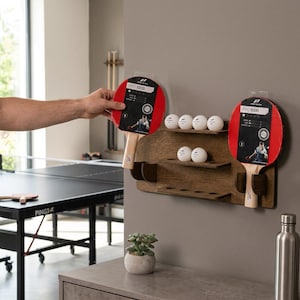 May include: A wall-mounted wooden rack with three shelves holding white table tennis balls and two red and black table tennis paddles labelled PRO 1000. The rack features circular cutouts for ball storage and slots for the paddle handles.