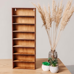 May include: A tall, rectangular wooden wall shelf featuring eight horizontal storage compartments with a natural brown wood grain finish and two circular mounting holes at the top for secure installation on a vertical surface.