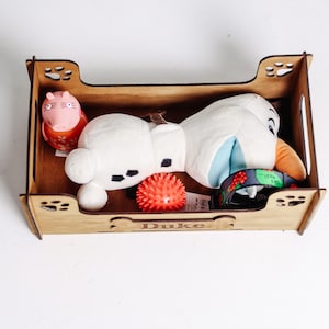 May include: A rectangular wooden pet toy box with paw print cutouts on the corners and the name Duke engraved on a bone-shaped plaque. Inside are a pink pig figure, a white plush snowman toy, a small red spiky ball, and a dark patterned collar.