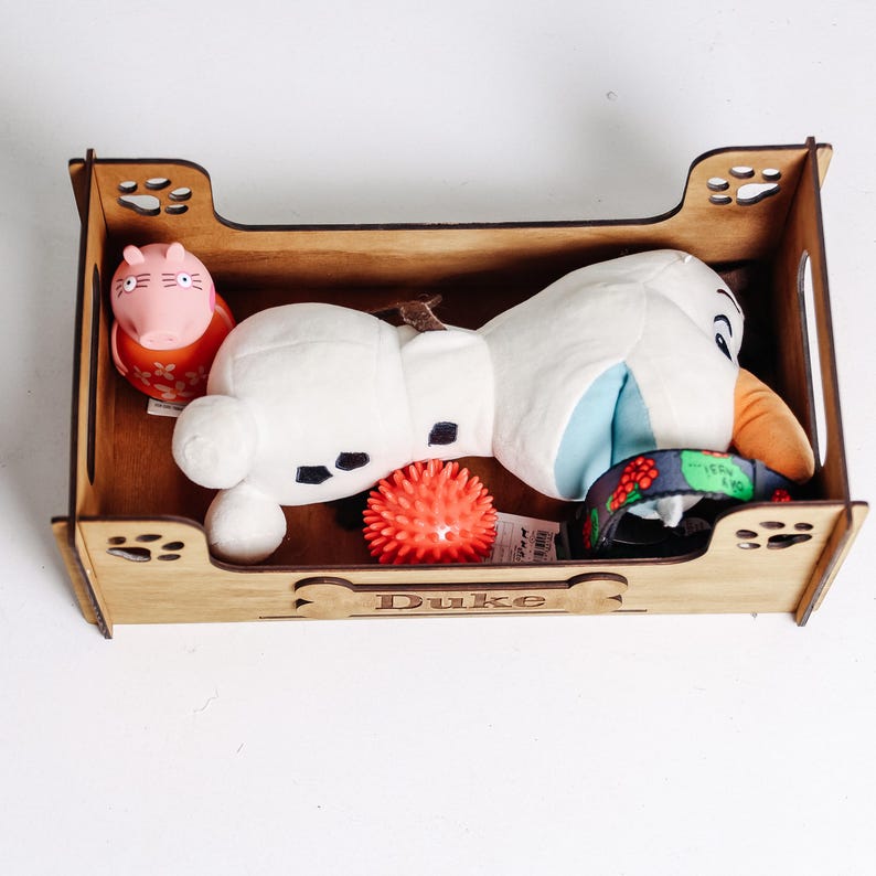 May include: A rectangular wooden pet toy box with paw print cutouts on the corners and the name Duke engraved on a bone-shaped plaque. Inside are a pink pig figure, a white plush snowman toy, a small red spiky ball, and a dark patterned collar.