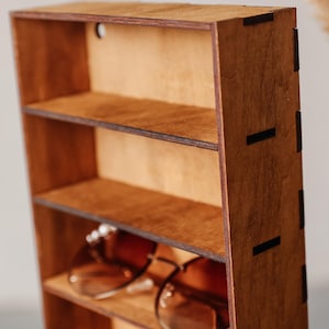 May include: A vertical wooden storage rack with four open shelves, constructed from dark brown plywood with visible interlocking joints on the side panel. A pair of brown-tinted spectacles rests on the bottom shelf of the wooden organiser.
