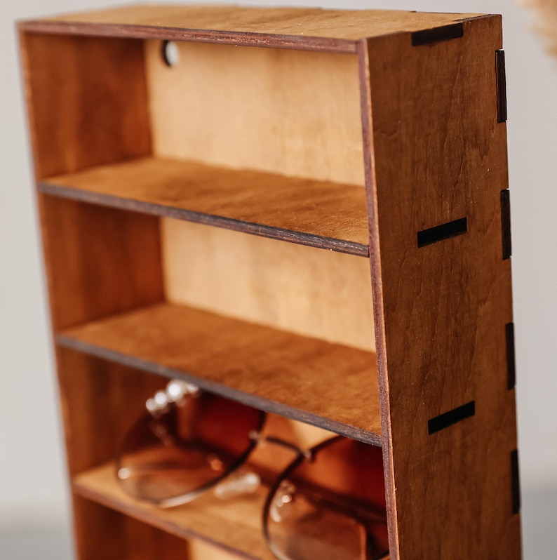 May include: A vertical wooden storage rack with four open shelves, constructed from dark brown plywood with visible interlocking joints on the side panel. A pair of brown-tinted spectacles rests on the bottom shelf of the wooden organiser.