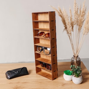 May include: A tall, rectangular wooden wall-mounted display shelf with seven horizontal compartments. The unit features a medium-brown wood grain finish and visible side joinery. It holds three pairs of spectacles on its shelves.