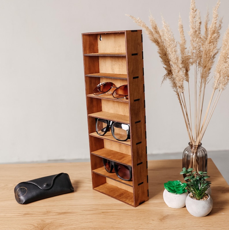 May include: A tall, rectangular wooden wall-mounted display shelf with seven horizontal compartments. The unit features a medium-brown wood grain finish and visible side joinery. It holds three pairs of spectacles on its shelves.
